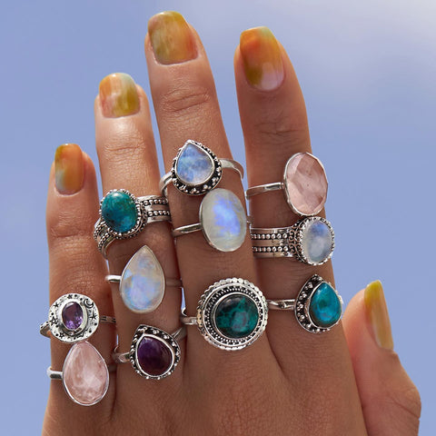 A close up image of a hand heavily adorned with precious stone silver rings, including chrysocolla, rose quartz, amethyst and moonstone on the background of a clear blue sky. The nails feature a water coloured - marble manicure that is coloured with orange, greens and blues. 