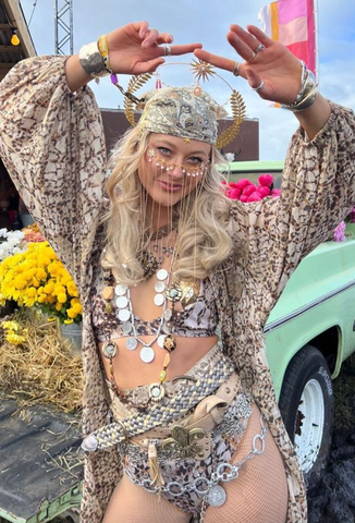 Dressed in a matching 3 piece brown and white snake skin set, a lady is mid dancing in a flowy shawl, bikini bottoms and a bikini top. She has accessorised with a matching head bandanna, chained coin necklaces and a thick wide similar patterned belt. She has a feathered halo head piece in gold, chains running down her cheeks from a necklace adjacent style jewel.  