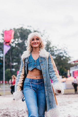 A young blonde woman in an all denim outfit, reminiscent of the 1970s. She sports a denim vest with low rise washed denim jeans and a fur trimmed denim coat, walking through a festival setting. 