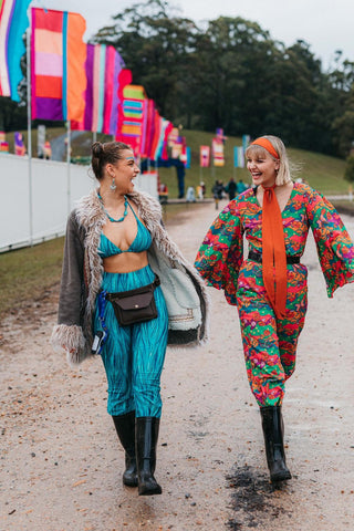 Two young women in colourful outfits smiling at each other as they walk through the muddy entrance at a festival. The woman on the left is wearing a matching turquoise 2 piece outfit, sporting a bikini top and long pants keeping warm in a brown fur trimmed and lined jacket. The lady on the right is wearing a colourful bright orange, pink and green jumpsuit, with flared long sleeves. Cinched in with a wide black belt, she has a matching orange head scarf. 