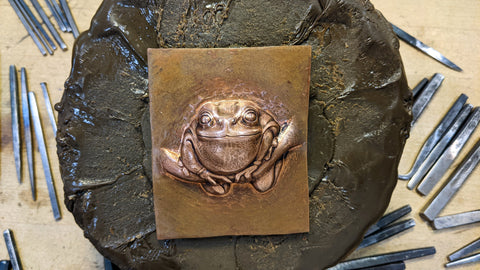 An image of a frog on a branch that has been raised in copper. The frog is chubby and delightful.