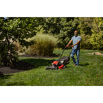 CRAFTSMAN M220 150cc 21-in. Self-propelled Gas Push Lawn Mower mowing near flowerbed with trees wearing jeans and blue shirt