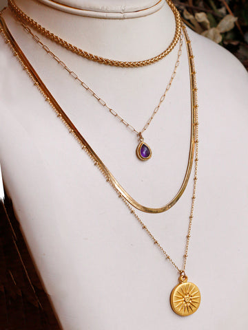 4 layered gold necklaces - a rope chain choker, a teardrop amethyst pendant necklace, a flat herringbone chain and a long gold coin pendant on a beaded gold chain.