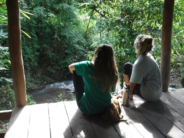 Two girls sitting on the edge of a dock with a dog overlooking a Thai river.