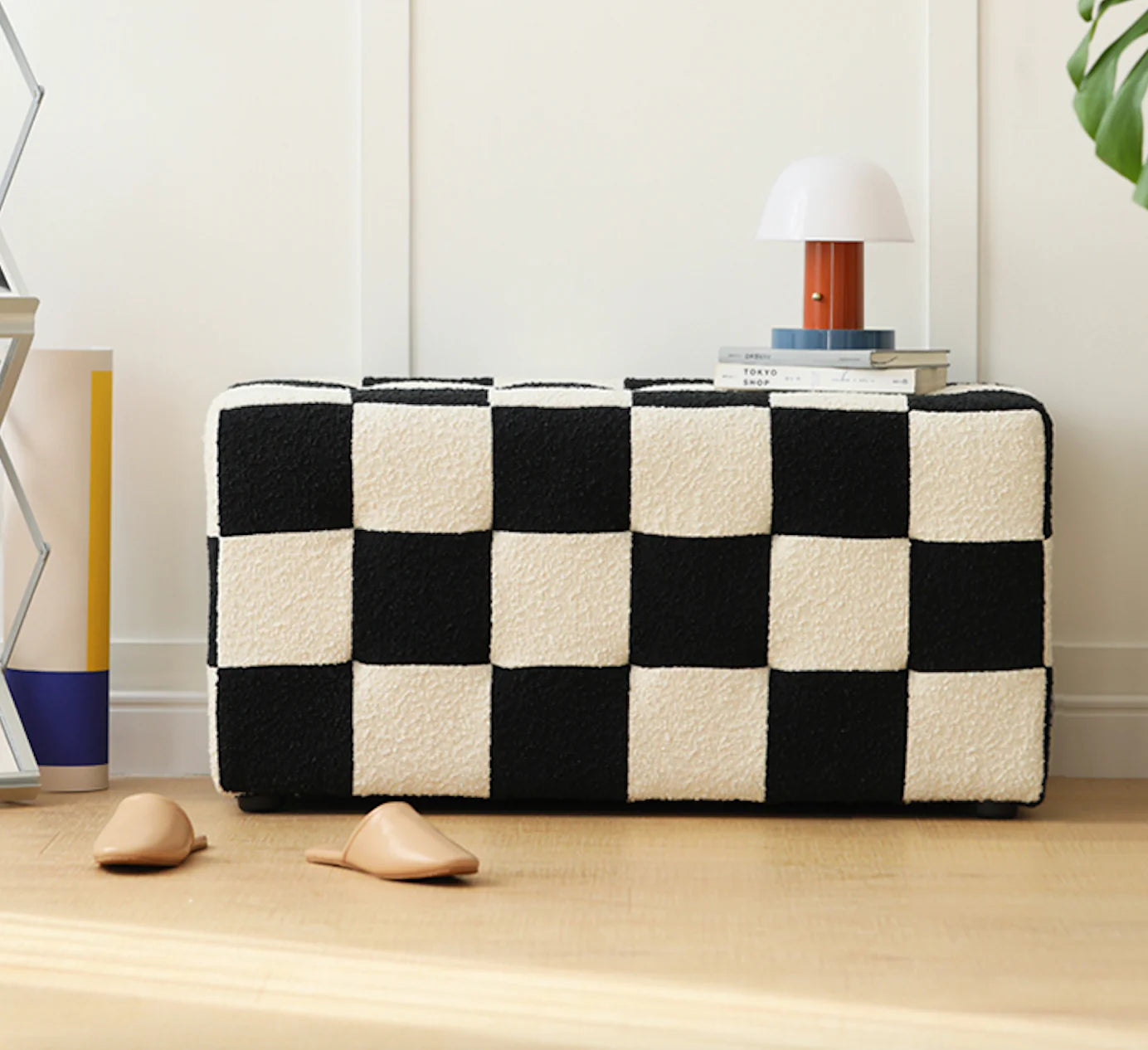Modern rectangular ottoman with a bold black and white checkerboard bouclé upholstery, placed on a wooden floor with decorative books, a lamp, and beige slippers nearby, creating a stylish and cosy interior setting.