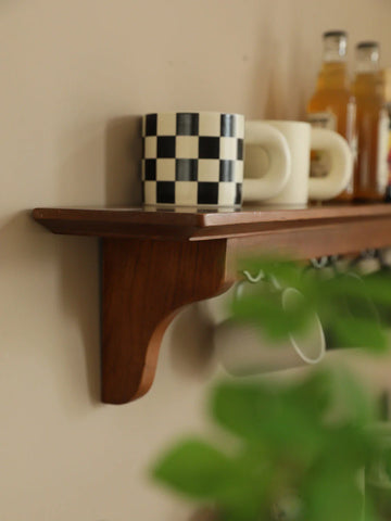 Close-up of a minimalist wooden wall shelf adorned with a black-and-white checkered mug, surrounded by blurred greenery and household items in the background.