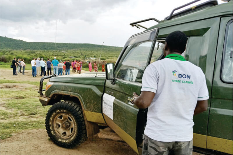 Guia do motorista do Bon Voyage Budget Safari no operador turístico do Quênia em pé perto do jipe verde observando de perto os turistas se divertindo com o povo Maasai