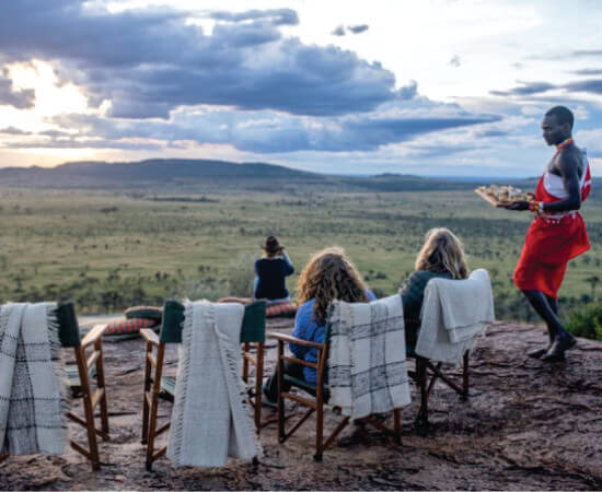 Hombre masai caminando cerca de tres personas disfrutando de una experiencia gastronómica única en un Sundowner en el lujoso tour grupal Mara Serena Safari Lodge Masai Mara