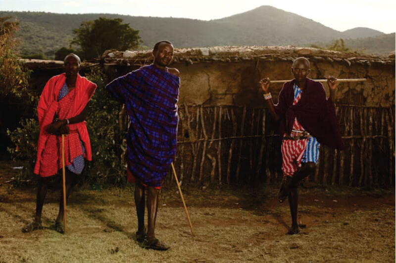 Drei Massai-Krieger stehen vor einer Lehmhütte im Masai-Dorf auf Massai-Dorf-Tour