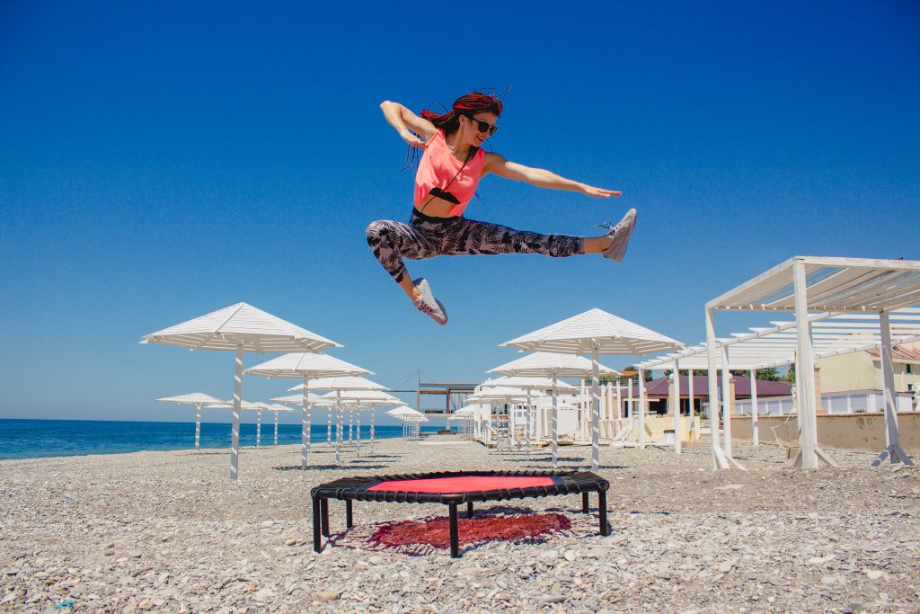 A slim athletic girl with dreadlocks jumping on a fitness trampoline outdoors on a pebble beach by the sea
