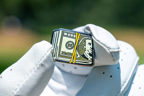 A hand wearing a golf glove holds up a golf ball marker that resembles a roll of money.