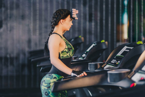 An image of a woman using a treadmill in a gym. She is wearing camouflage print sportswear and adjusting her headphones. The background is blurred, but visible equipment and mirrors suggest the context of a fitness center. The overall scene evokes a sense of activity and focus