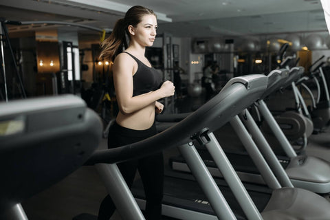 A woman exercising on a treadmill in a modern and well-equipped gym. She wears a black sports top and tight pants, her hair tied in a ponytail. Her gaze is focused ahead as she runs steadily on the treadmill. The treadmill is positioned in a spacious room with gray walls and a floor resembling marble or similar material.