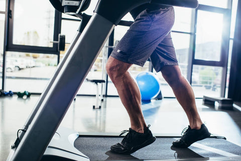 A photograph of a corner inside a gym. A person wearing gray shorts and black sneakers is exercising on a treadmill located slightly to the left of the center of the frame. In the background, other fitness equipment such as dumbbells and other workout machines can be seen.