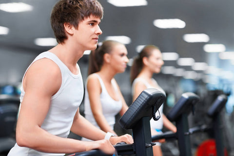 A photo showing three individuals using treadmills in a modern and well-lit gym. In the foreground, a young man in a white vest and dark shorts is running attentively on a treadmill. He has brown short hair and appears focused. In the background, two women are also exercising on separate treadmills. The woman on the left wears a white sleeveless top and light-colored pants, while the woman on the right is dressed in a white top and black pants.