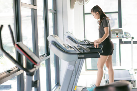 A photograph of a woman using a treadmill in a gym. She wears a black top and shorts, paired with blue sneakers. Her hair is tied in a ponytail, and she is adjusting the settings on the treadmill. In the background, other fitness equipment such as dumbbell racks and exercise bikes can be seen. Natural light filters through the windows, creating a well-lit and inviting atmosphere.