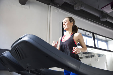 A woman exercising on a treadmill in a gym. She wears a black sports bra and blue leggings, focusing her gaze forward. The treadmill is black with gray trim, and in the background, windows and exhaust vents on the ceiling can be seen.