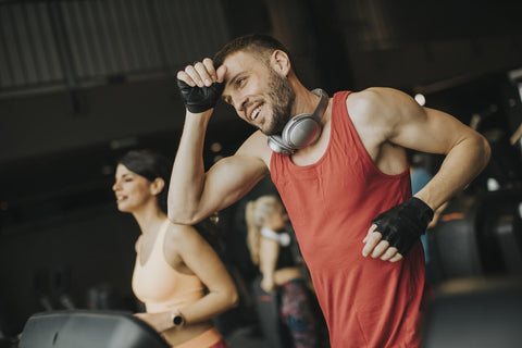 A scene inside a gym featuring a man in a red tank top and black gloves. He wears headphones and performs a stretching exercise, likely to relax after a workout. His expression is relaxed, smiling as he glances to the side. Behind him, a woman can be seen using a treadmill, dressed in an orange top with her hair tied in a ponytail, focused on the distance ahead.