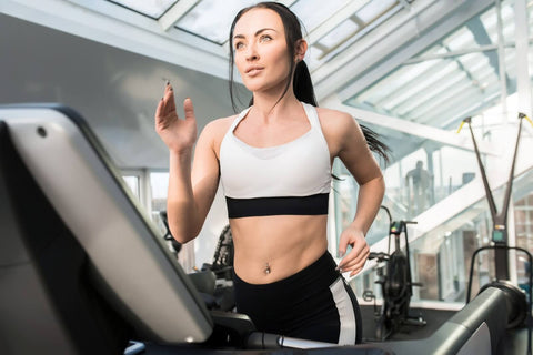 A woman is exercising on a treadmill inside a gym. She is wearing a white sports bra and black sports shorts, with her hair tied in a ponytail. She is looking forward with a focused expression, and her body is slightly leaning forward in a dynamic posture while exercising. The treadmill is located indoors, and in the background are glass windows and metal structures.