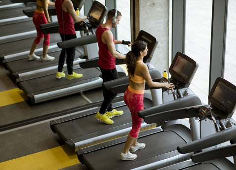 A photograph of a corner of a gym, showcasing three individuals exercising on treadmills. The person on the left wears a red workout suit and utilizes a black treadmill, while the middle individual sports an orange and yellow ensemble on a similar model. The person on the right, dressed in pink, also uses the same treadmill. All three seem engaged in aerobic activity.