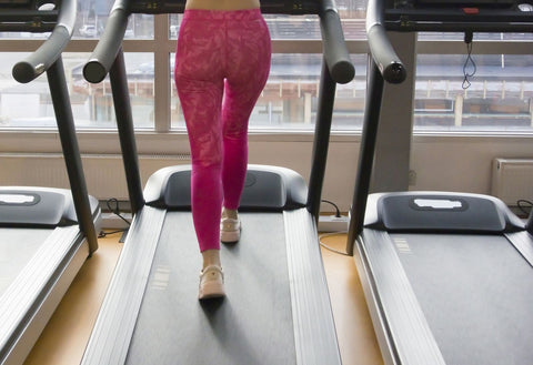 A woman exercising on a treadmill in a modern and well-lit gym. She is dressed in pink tights and light-colored sneakers, with her back to the camera. The treadmill is placed in a room with large windows that showcase the buildings outside. The walls are painted white, contributing to a clean and sleek appearance.