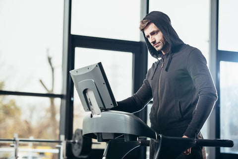 A photo depicting a male wearing a black hoodie standing inside a gym. He is intently focused on a screen attached to a treadmill. The treadmill is located in a spacious room that features large windows and various fitness equipment. The male stands close to one side of the treadmill, his right hand resting on the handrail.