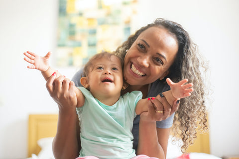 The picture is of a young girl, who appears to be very happy and is smiling cheekily. She is holding her hands outstretched, up in the air. The child has wavy brown hair. There is an adult behaind the child, holding the childs arms to keep her balanced. She has curly brown hair and is smiling broadly. SUNNYBOD™ Refillable Sunscreen Brush applicator.