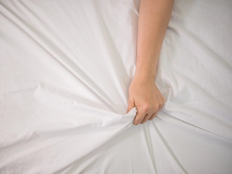 Mano femenina sujetando sábana blanca, expresión de placer íntimo al aprender vibrador como usar en pareja.