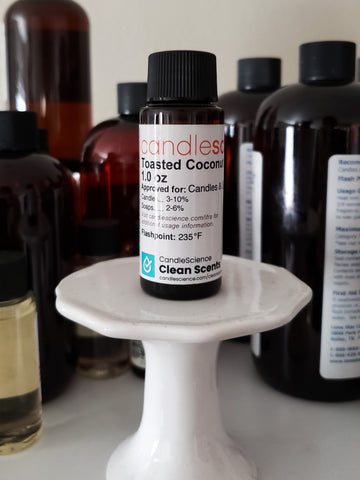 Small brown bottle with a white label with black text that says Toasted Coconut from CandleScience, sitting on a small white pedestal in front of black brown bottles of fragrance oil.