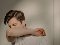 Caucasian male in a white tshirt standing in front of a shadowy, white background sneezing into the crook of his elbow. 