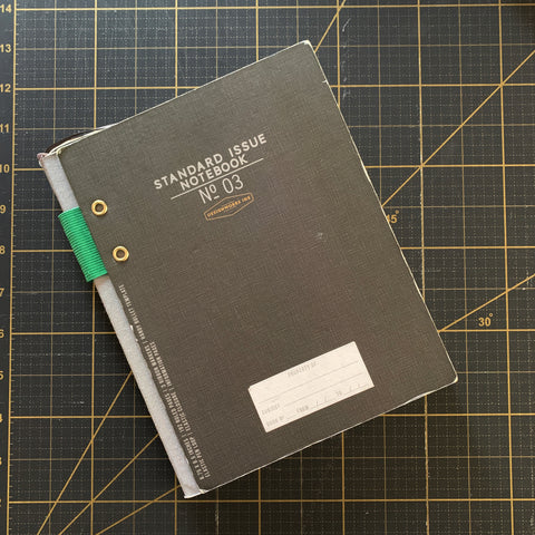 image of closed standard issue notebook from designworks ink on a black grid background