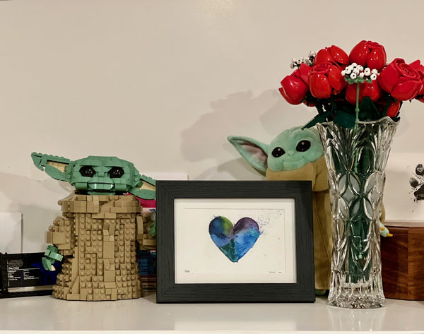 Lego Grogu next to a framed heart painting and next to Lego roses on a shelf