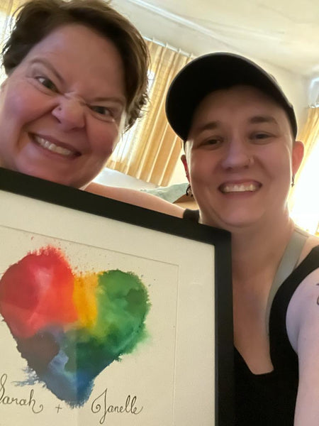 Janelle and Sarah, two white women, smiling and holding up their framed rainbow heart painting with their names in calligraphy