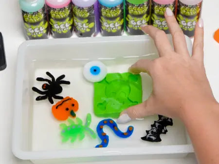 A hand arranges colorful gelatinous Halloween shapes in a water tray with gel paint bottles in the background.