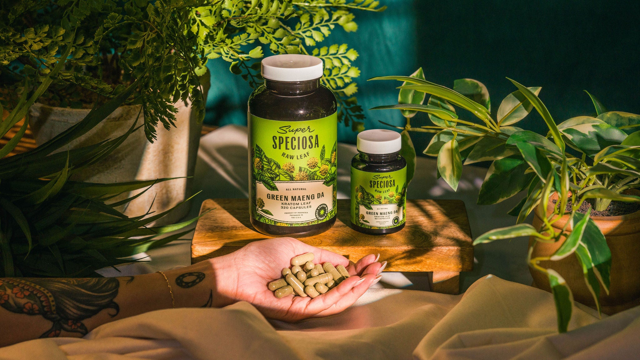 Person holding capsules near bottles of Green Maeng Da Kratom amidst lush green plants.