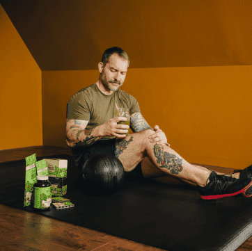 Man with tattoos sits on exercise mat, holding a drink, fitness products nearby.