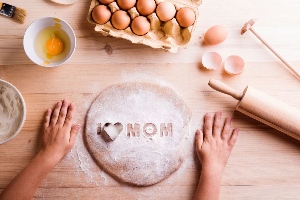 Child's hands on a dough with I LOVE MOM imprint, baking ingredients around.