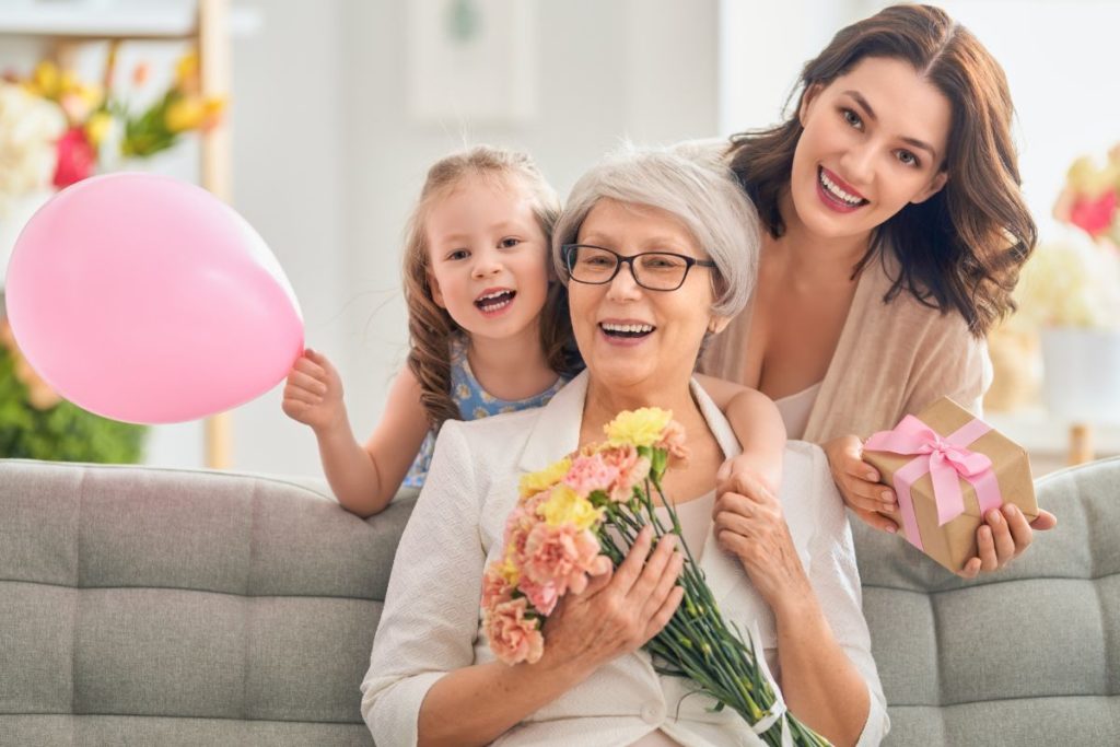 Three generations of women smiling; a child with a balloon, a grandmother with flowers, and a mother with a gift.