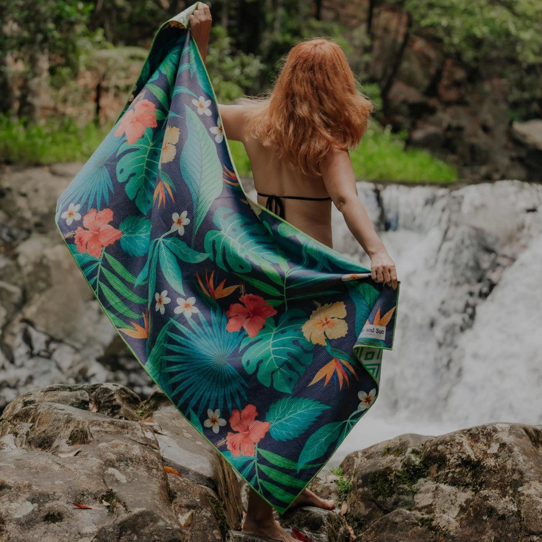 Woman standing in front of a waterfall, holding a tropical pattern towel stretched behind her.