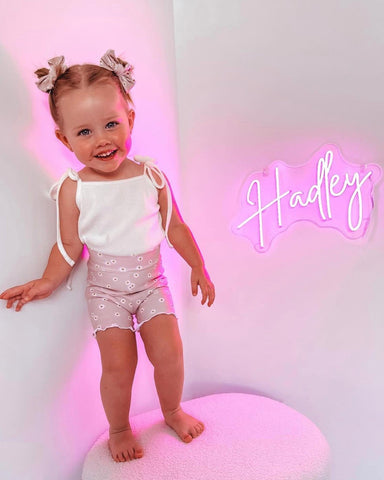 Little toddler girl with bows her in hair standing up on a fluffy stool smiling, against a white wall. Her bedroom wall has a pink neon sign of her name that says Hadley