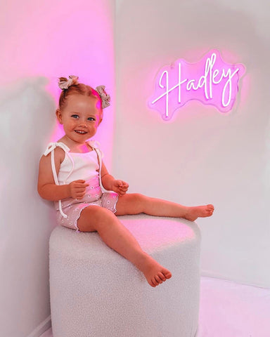 Little girl with bows in her hair sitting on a stool smiling and laughing, against a white wall with a pink neon sign of her personalised name