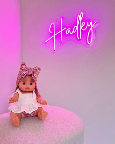 A little girl doll is sitting on a white fluffy stool with a bow in her hair, in a white bedroom. On the white bedroom wall is a hot pink neon sign that says Hadley.