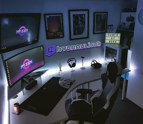 "A stylish computer desk setup for gaming, featuring a neon Twitch sign in the background, wall-mounted photos, a comfortable gaming chair, and a keyboard at the ready. The desk is well-equipped with its gamer neon sign