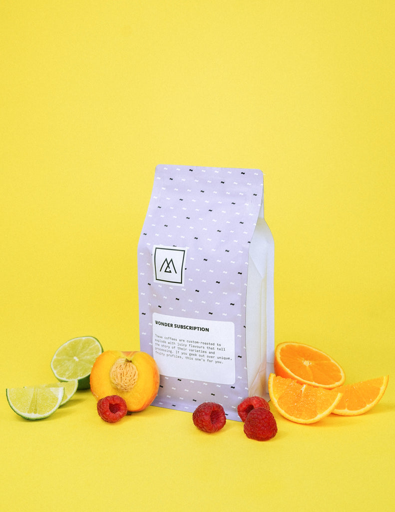 A purple bag of Monogram coffee sits on a yellow background and is surrounded by limes, peaches, raspberries and oranges.