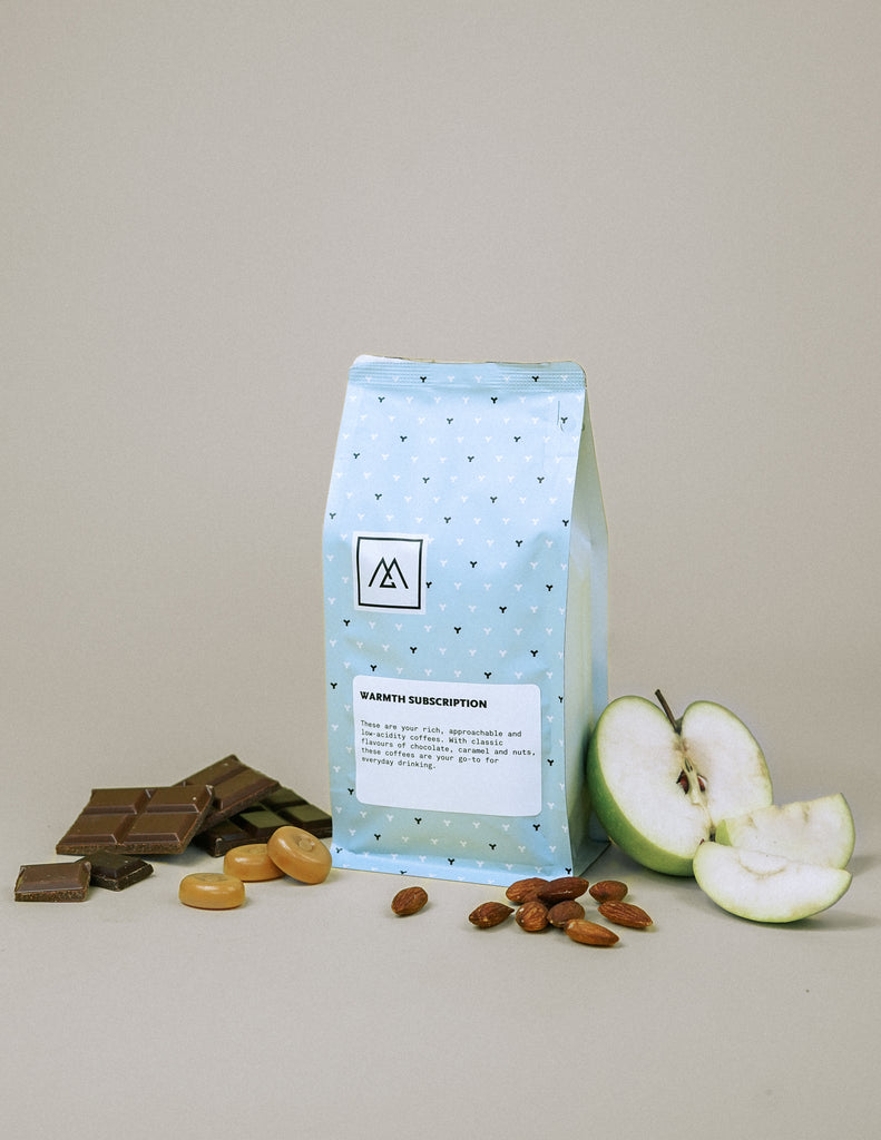 A blue bag of Monogram coffee sits on a grey background surrounded by chocolate, caramel, nuts and green apple.