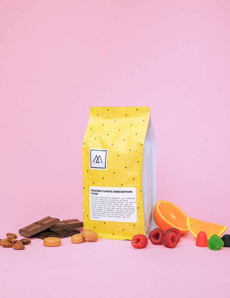 A yellow bag of Monogram coffee sits on a pink background and is surrounded by chocolate, nuts, caramel, raspberries, oranges and jube jubes.