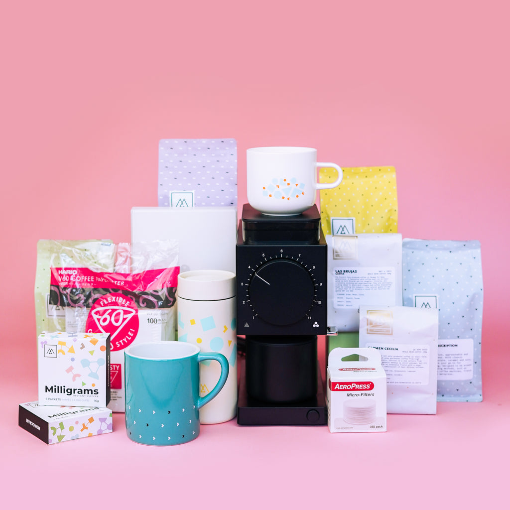 An array of products from Monogram coffee sit on a pink background. Included in the photo are Milligrams instant coffee, V60 filters, a Fellow Ode Grinder, a Loveramics mug, a Miir travel tumbler