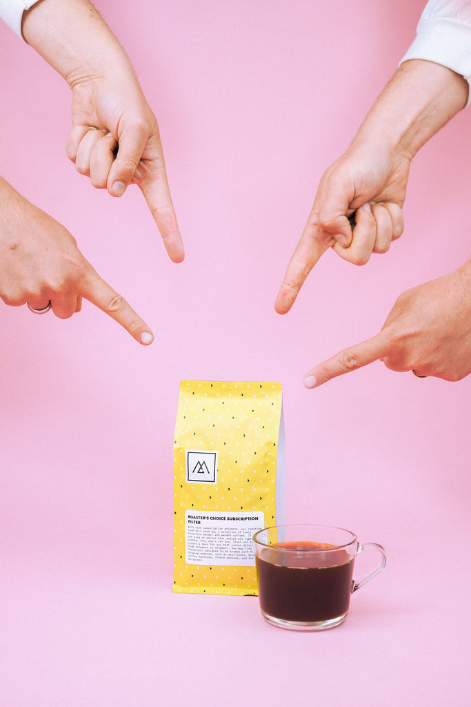 A yellow bag of Monogram coffee sits on a pink background with a cup of brewed coffee beside it and 4 fingers pointing to the bag of coffee.