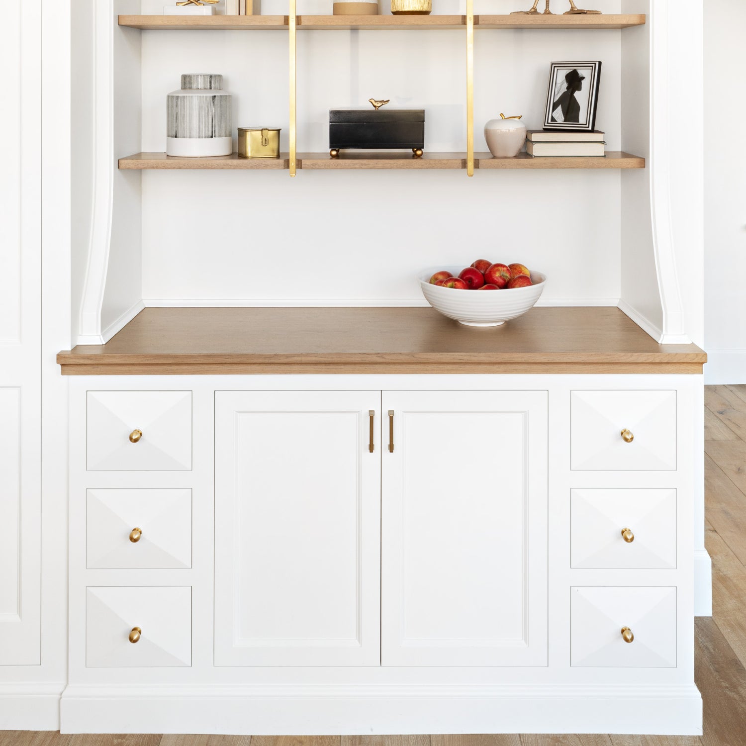 Elegant white sideboard cabinet with gold handles, set below wooden shelves adorned with chic home decor.