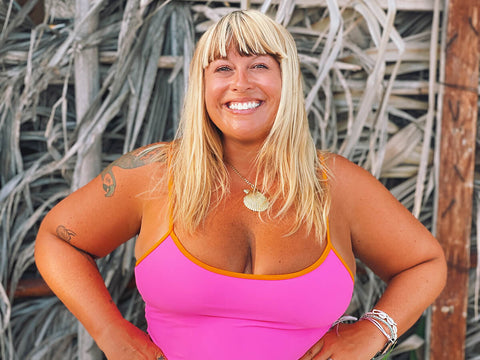 Heather Brown, a blonde haired woman with bangs, wears a pink swimsuit and smiles at the camera with her hands on her hips.